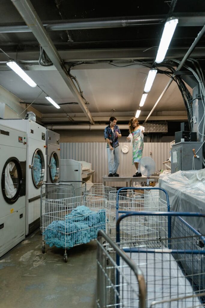 pexels-photo-8774385 Two young adults examine equipment in a commercial laundry facility, surrounded by washing machines and carts.